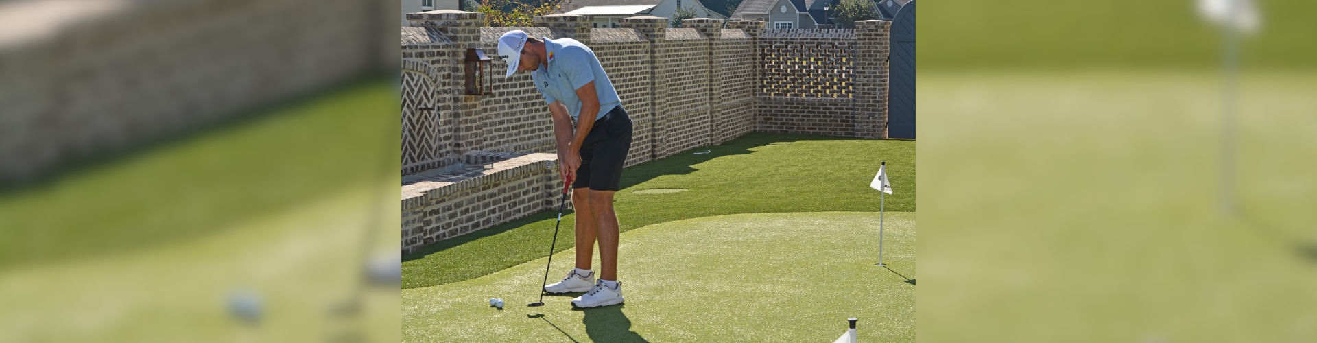 Golfer practicing putting on a backyard putting green with a flag pin, surrounded by a decorative brick privacy wall in a residential neighborhood