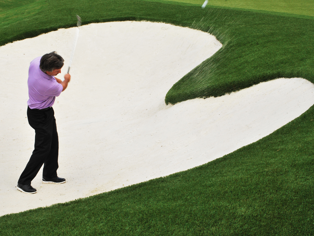 Golfer hitting a bunker shot beside a professionally designed backyard putting green with realistic sand trap and manicured artificial turf.