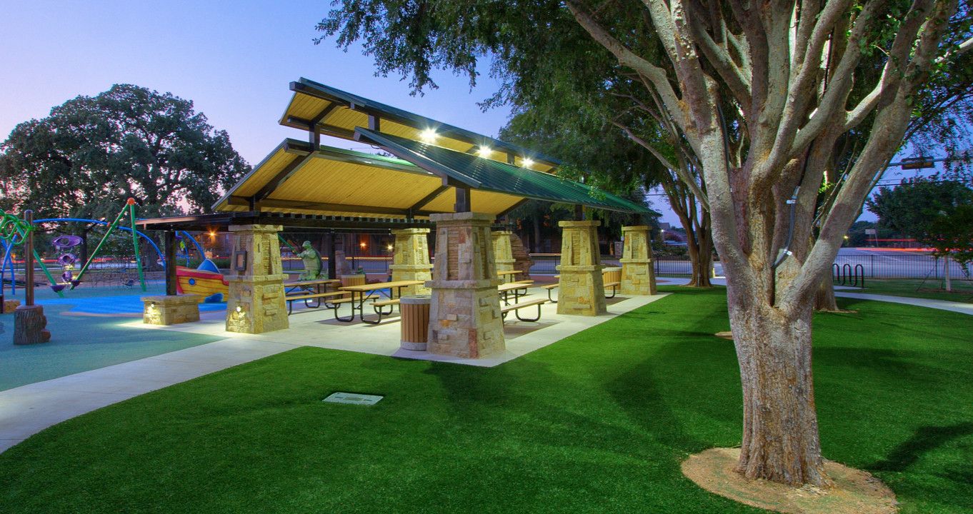 Modern public park pavilion at dusk featuring lush green lawn from professional synthetic turf installation, stone columns, picnic tables, and a shaded playground surrounded by mature trees.
