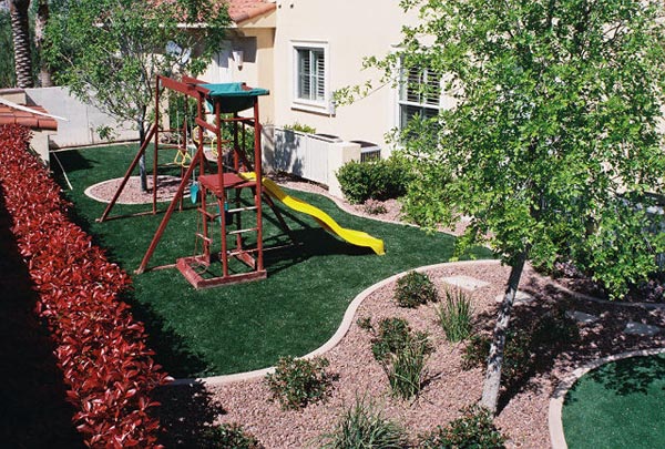 Fake grass installation in backyard play area with wooden swing set, slide, and desert landscape border plantings