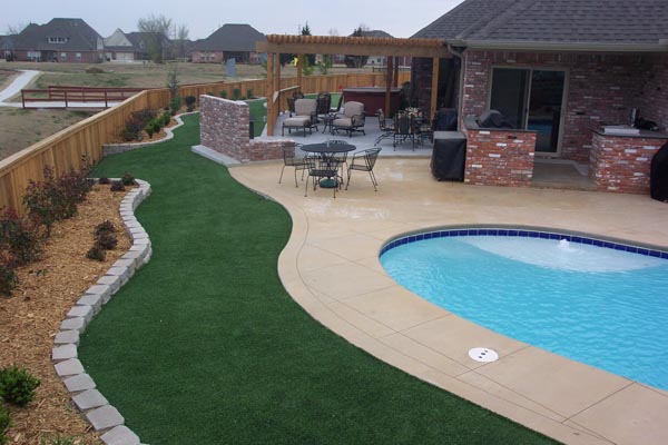 Fake grass installation alongside residential pool and patio with pergola, outdoor seating, and stone border edging