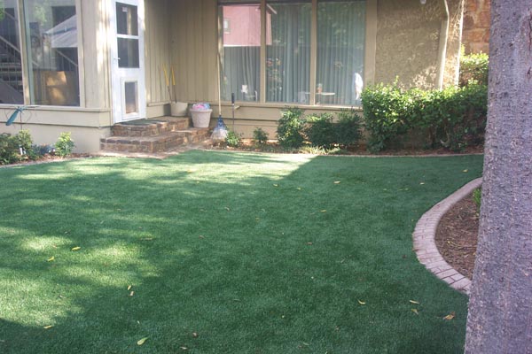 Synthetic grass installation in residential backyard with brick border edging, garden beds, and mature shade trees