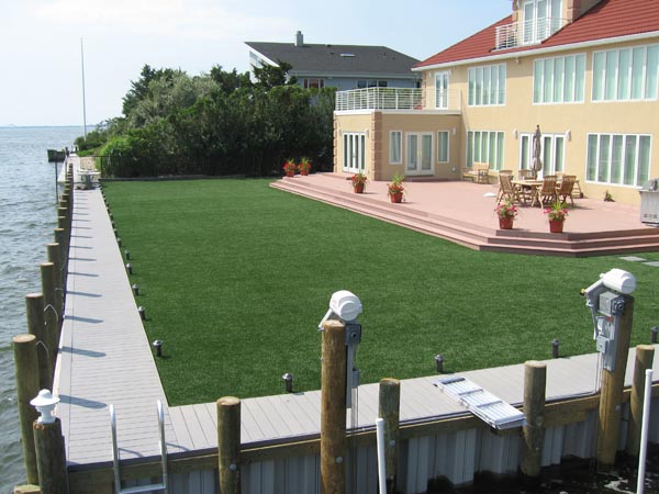 Synthetic grass installation between waterfront dock and luxury home patio with outdoor dining area and water views