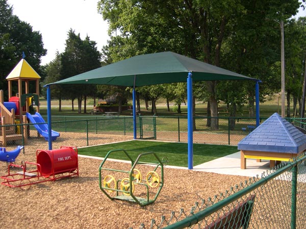Synthetic grass installation under shaded canopy structure at public park playground with colorful play equipment