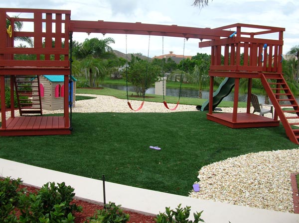 Synthetic grass installation in backyard play area with large red wood swing set, slide, and decorative gravel borders