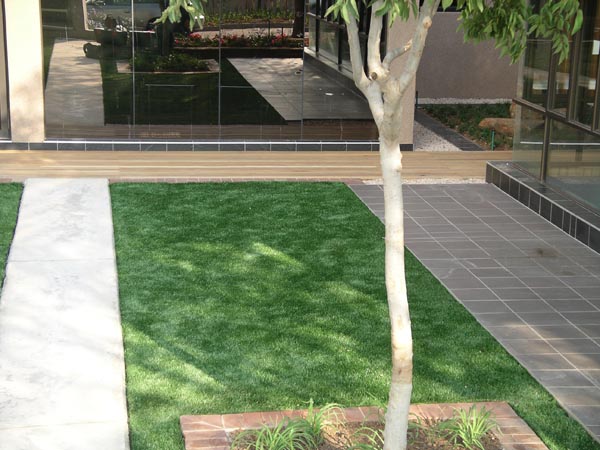 Synthetic grass installation in modern commercial office building courtyard with tile walkways and shade tree