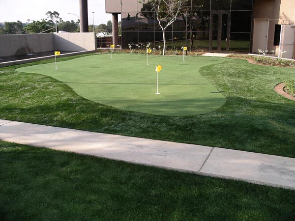 Synthetic grass installation with multi-hole putting green and yellow flags at corporate office building exterior