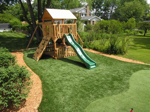 Artificial grass play area with wooden swing set and slide surrounded by mulch border and lush backyard landscaping