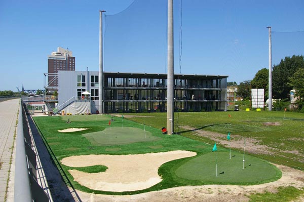 Outdoor putting green with multiple flags and sand bunkers at urban commercial golf training facility with driving range nets