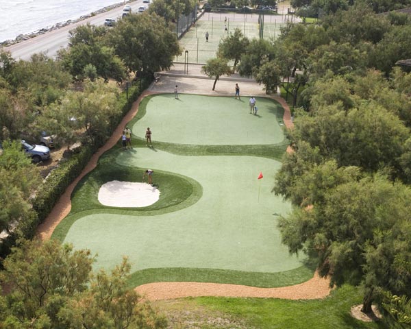 Aerial view of outdoor putting green with sand bunker and red flag at coastal resort property surrounded by trees and tennis courts