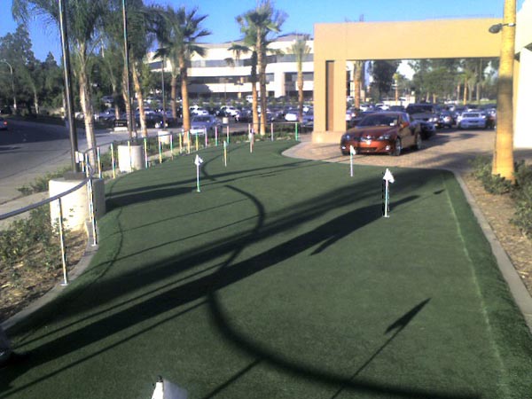 Commercial artificial grass putting green with multiple flags and palm tree shadows at corporate office complex parking area