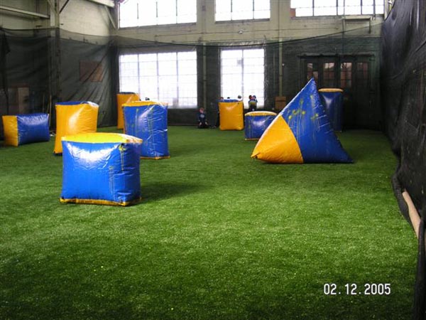 Indoor artificial turf paintball arena with blue and yellow inflatable obstacles inside large warehouse facility