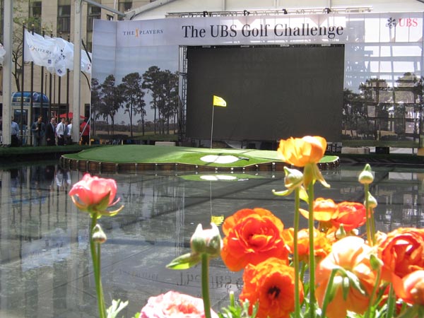 Artificial grass putting green display at UBS Golf Challenge corporate event with yellow flag and colorful flowers in foreground