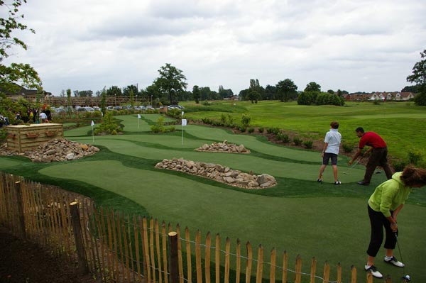 Group of golfers playing on artificial turf putting green with rock landscaping and wooden fence surround at outdoor golf facility