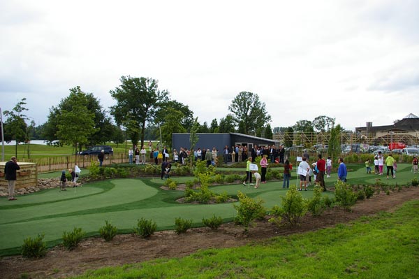 Crowd of visitors gathered around artificial turf putting green at public park grand opening event with lush landscaping