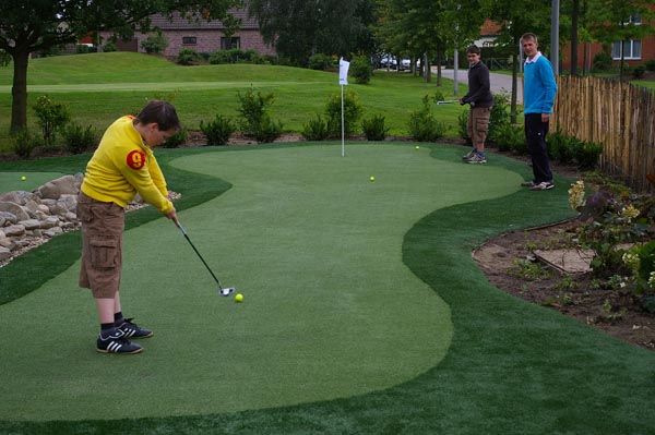 Child and adults playing on synthetic turf putting green with rock border and garden landscaping in residential backyard