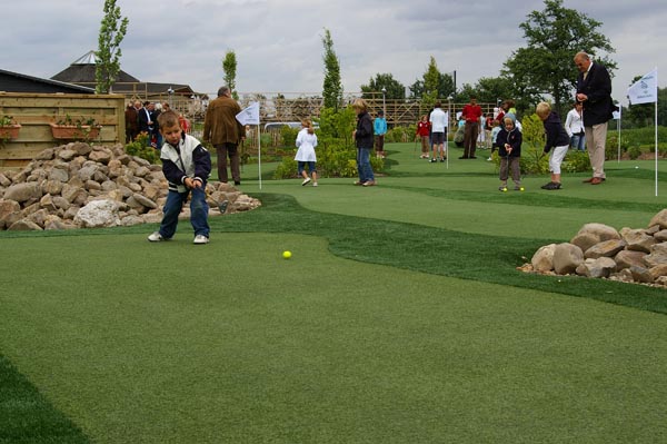 Child putting on artificial putting green with boulder landscaping and crowd of spectators at outdoor golf event