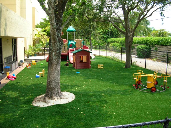 Artificial turf installation at commercial daycare or school playground with play structures, toys, and mature shade trees