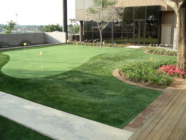 Synthetic turf putting green with yellow flags and colorful flower beds at corporate office building exterior