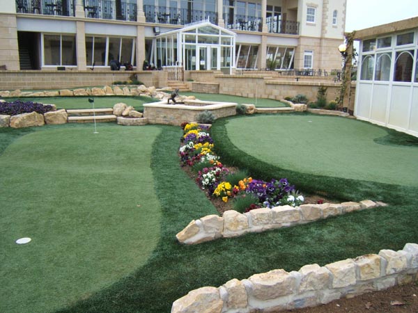 Synthetic turf putting green with stone retaining walls and flower bed landscaping at luxury hotel or resort property