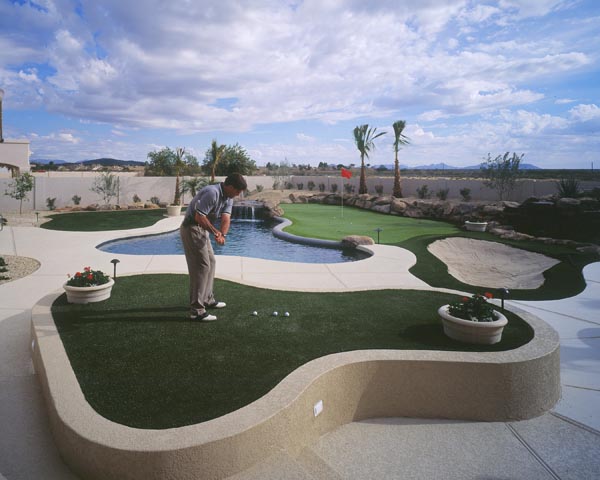 Golfer practicing on backyard putting green with pool, waterfall feature, sand bunkers, and palm trees in Southwest desert backyard