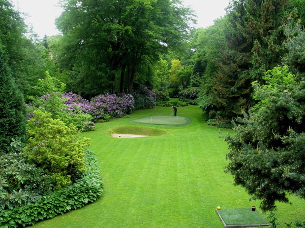 Golfer practicing on backyard putting greens with sand bunker set within lush estate garden of mature trees and flowering shrubs