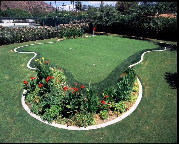 Backyard putting greens with red flag surrounded by curved colorful flower bed border and manicured lawn