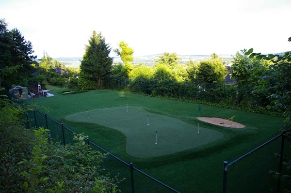 Backyard putting green turf with multiple flags, sand bunker, and panoramic valley views behind black metal fence