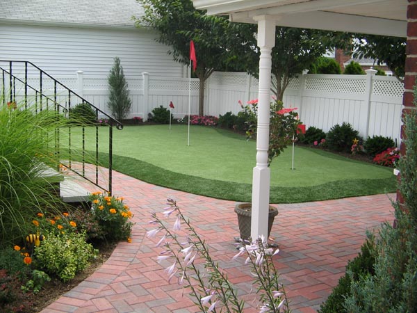 Backyard putting greens with red flags and flower bed borders next to brick paver patio and white vinyl fence