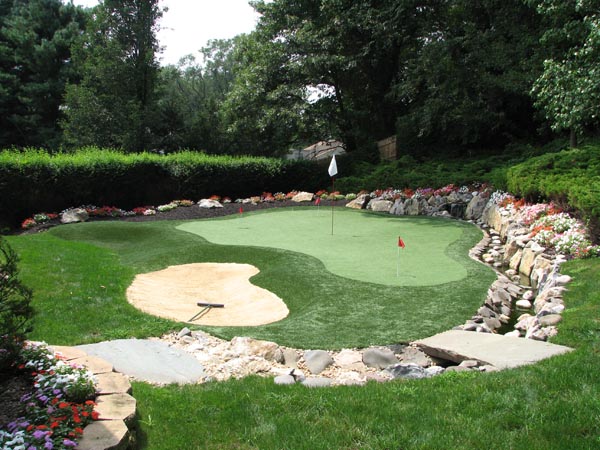 Backyard putting greens with sand bunker and rock border landscaping surrounded by colorful flower beds and mature trees
